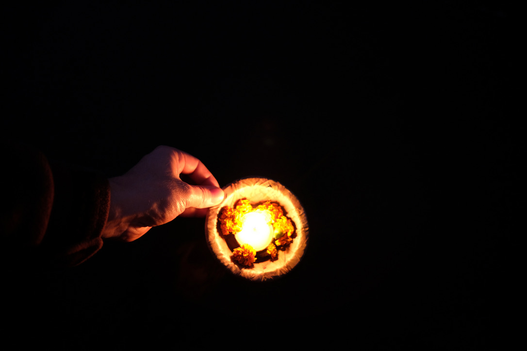 A hand holds a burning oil lamp in a small bowl made of leaves against a deep black background. The flame is surrounded by orange blossoms and casts a warm, bright light on the fingers.