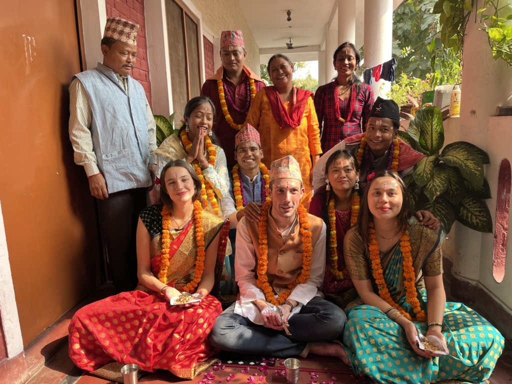 Group photo of my smiling neighbors on our half-open veranda. In the foreground, three young volunteers are sitting cross-legged on the floor. They are wearing festive clothing, orange flower garlands around their necks, and red dots on their foreheads. Behind them, other people are standing and sitting, some of whom are wearing patterned traditional Nepalese headgear.