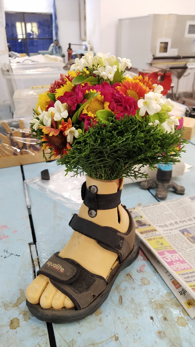 Color close-up: A bouquet of colorful flowers and conifer greens stuck in a prosthetic foot shell. The skin-colored prosthesis is wearing a brown Velcro sandal. The background is an untidy workbench.