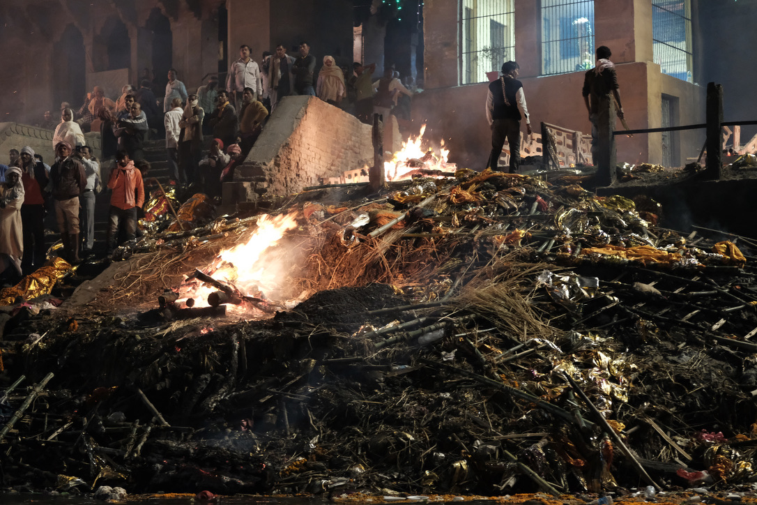 Night scene at a cremation ghat with burning wood fires in the foreground and devout spectators on stone steps in the background.